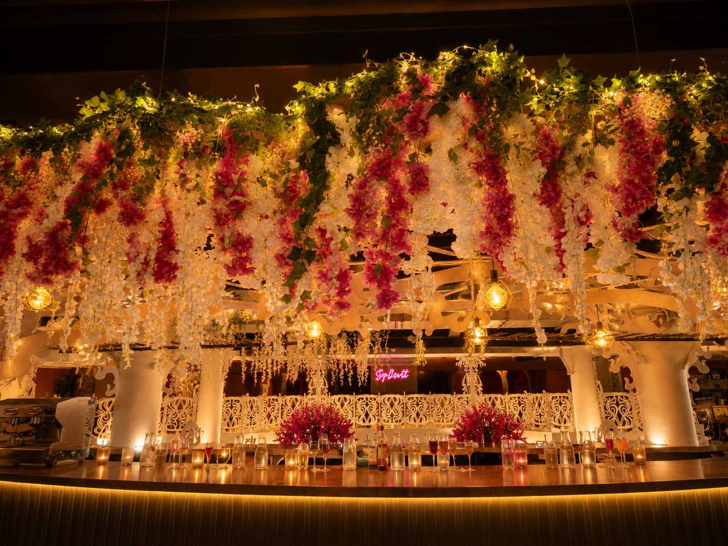Curved bar at Opulent @ 35 Liverpool lit by warm pendant lights with cascading pink and white blossom canopy overhead and gold-rim mocktail flutes on the counter